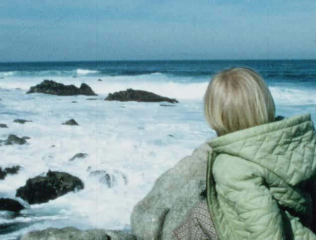 A white boy in a coat sits on rocks on the beach