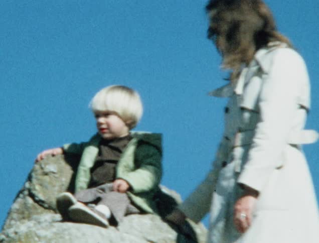 A woman standing beside her son on a tall rock formation as he sits and waves at the camera