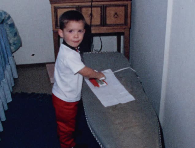 Male toddler uses a toy iron to pretend to iron a piece of cloth placed on an ironing board that is adjusted to his height