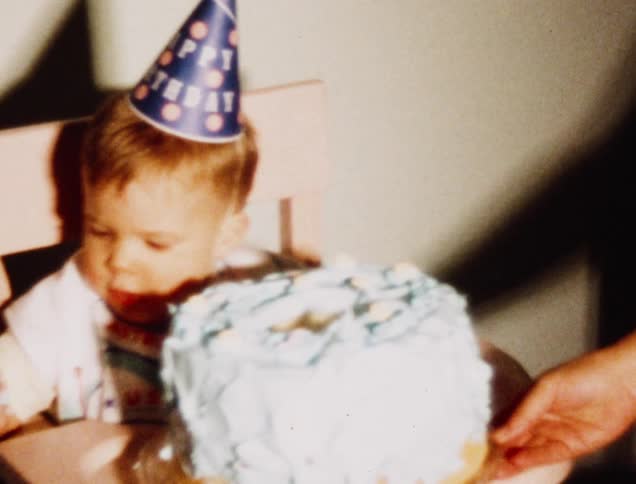 Birthday cake is placed in front of a baby wearing a party hat
