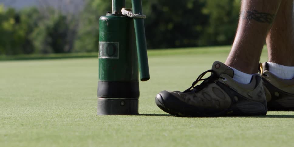 A man wearing athletic shoes uses a machine to create a hole on a putting green