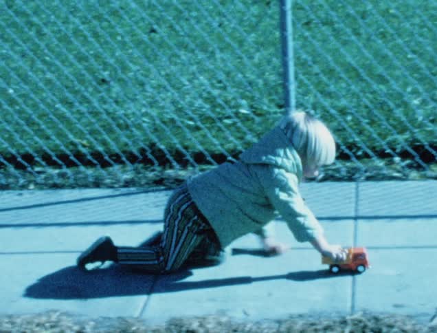 Small boy crawls on the sidewalk along a fence while pushing a toy truck