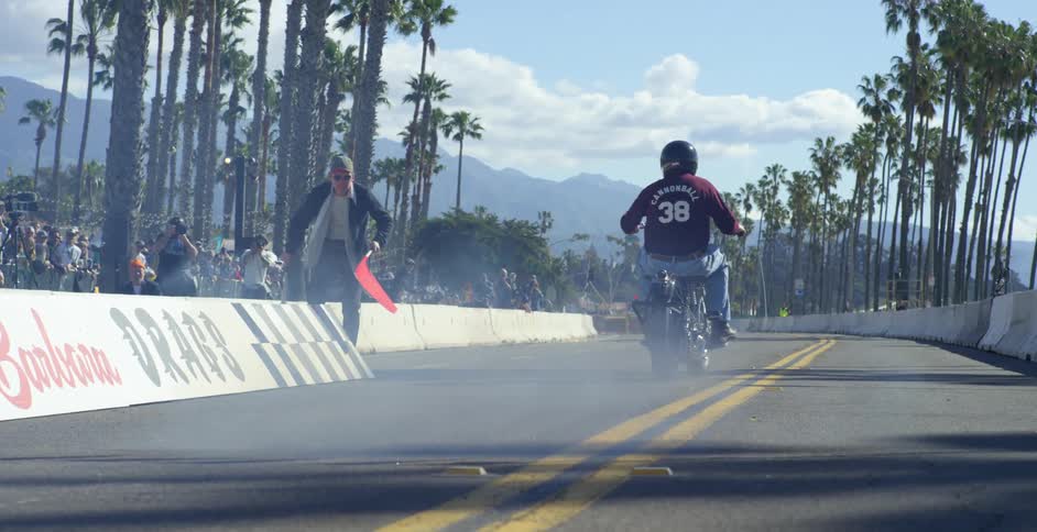 A crowd watches as a man jumps up and lowers the flag at the start of a motorcycle race
