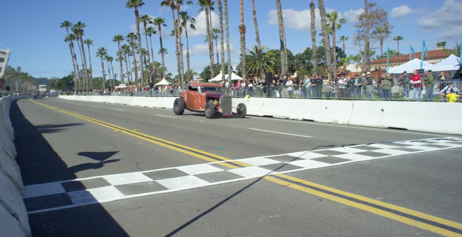 A classic race car crosses the finish line at an outdoor racing event with a crowd of spectators watching