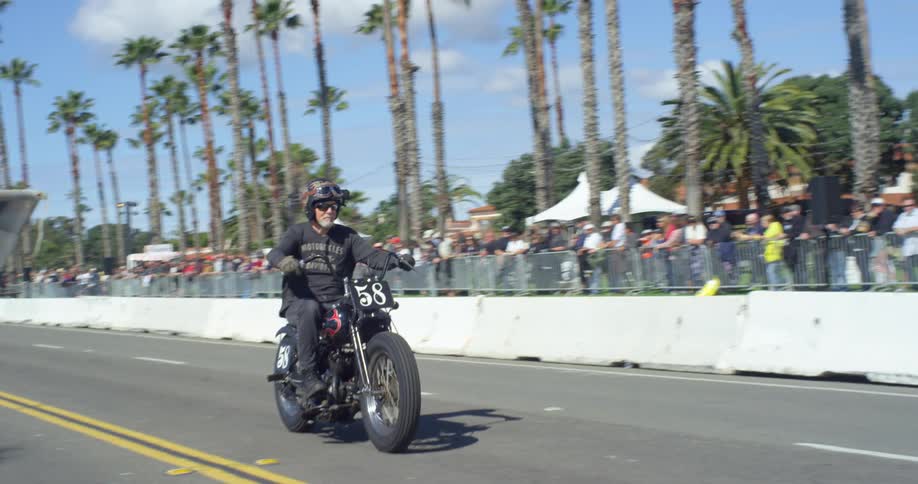 A man waves a checkered flag as an old white man rides by on a motorcycle