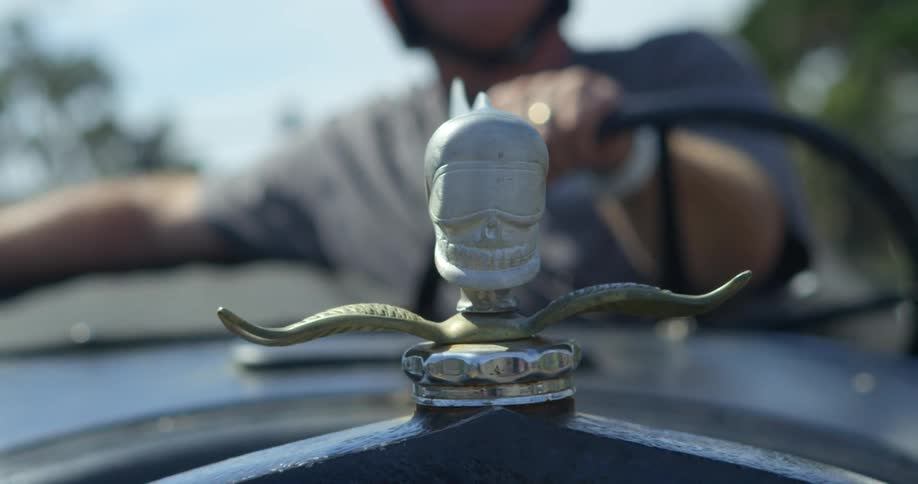 A man drives a classic car with a skull as the hood ornament