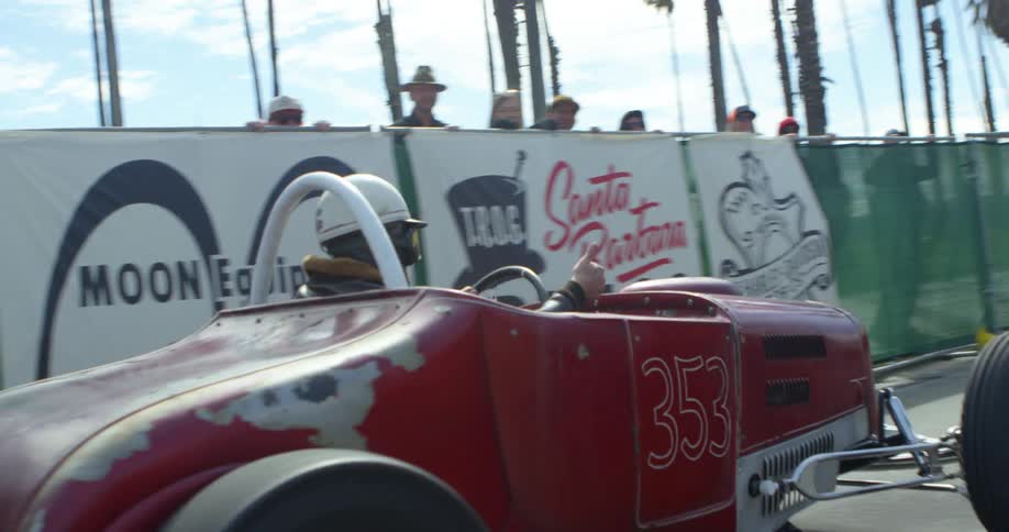 A man in retro clothing drves a vintage race car down a street behind other vintage vehicles as he points and gives a thumbs up to spectators