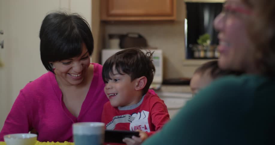 Women sit around table in kitchen welcome boy showing his tablet