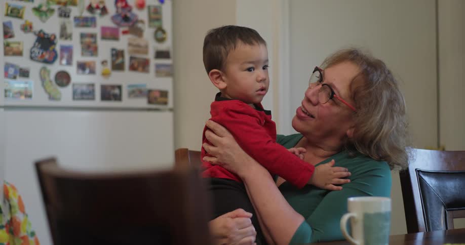 A woman seated in a kitchen holding a young boy smiles and kisses him on his cheek