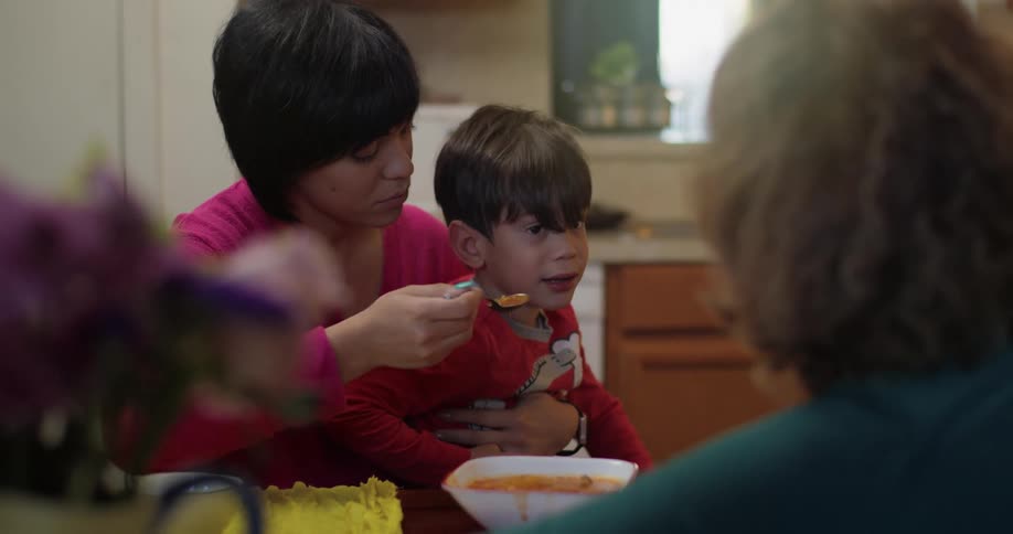 A mother tries to feed her distracted son at the kitchen table until he gets down and walks away