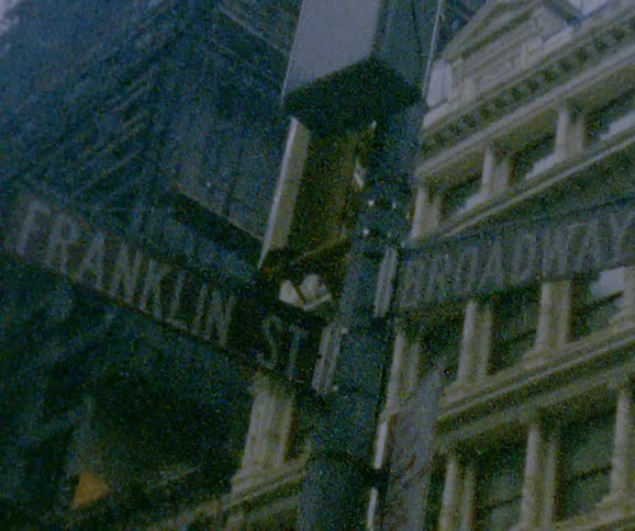 A sign post at the intersection of broadway and franklin with city buildings behind