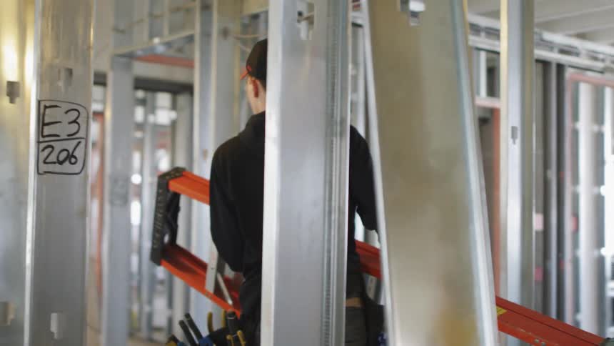 A young white man carries a ladder through a construction site