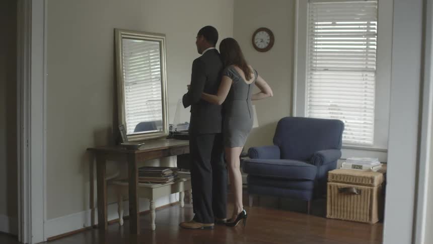 A man dressed in a business suit standing in front of a mirror is embraced by a woman
