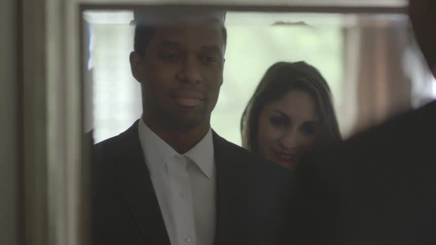 A man stand in front of a mirror adjusting his suit and a woman embraces him
