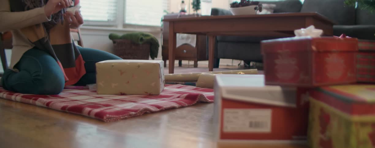 A woman kneels on a folded up blanket on the living room floor wrapping a box