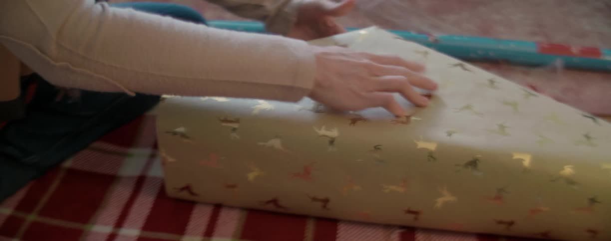 Female adjusts a box on top of christmas wrapping paper with gold reindeer and then folds the paper over the top of the box to wrap the gift