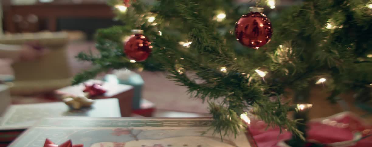 A christmas tree with christmas presents underneath with a person cutting wrapping paper in the background