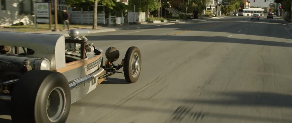 A project roadster with exposed engine and fat tires drives down a neighborhood street on a sunny day
