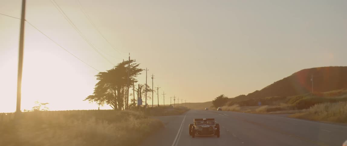 A small retro vehicle traveling down a countryside road alongside modern vehicles