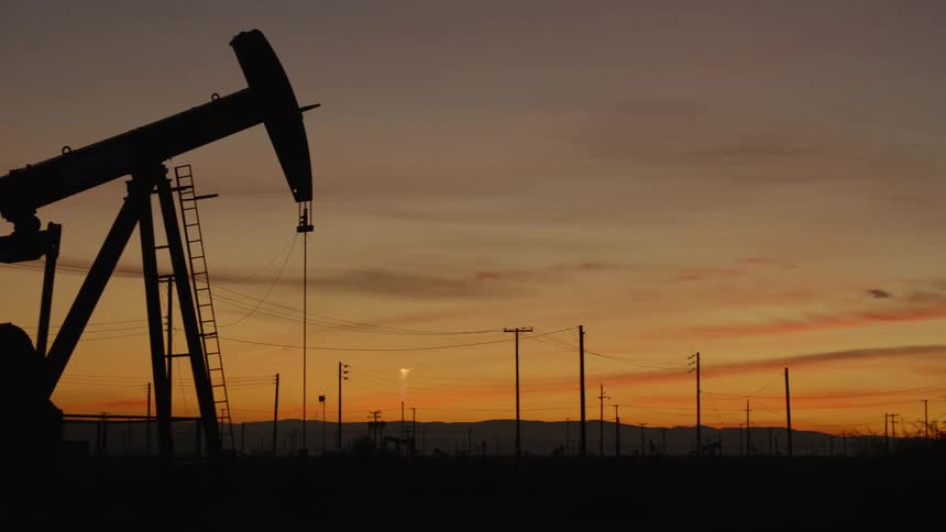 Oil pumps seesaw in plain crossed by power lines with mountains on horizon under orange sky