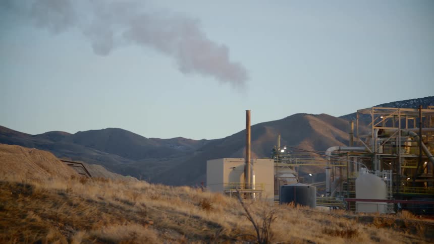 A refinery with a chimney with smoke with mountains and the sky in the background