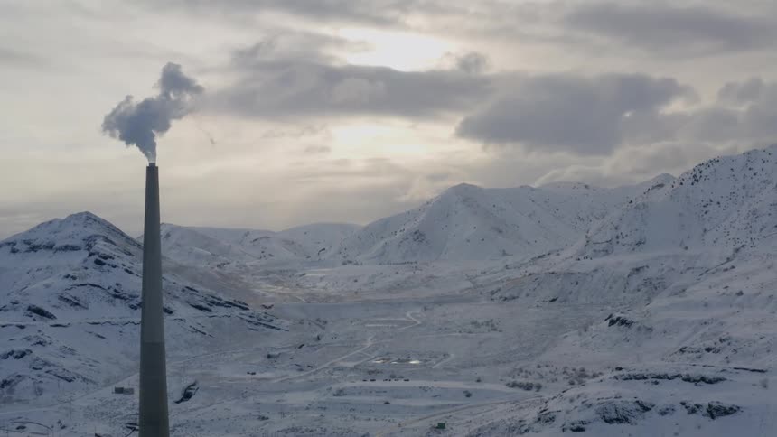 Tall chimney releases smoke into air in snow covered valley surrounded by snowy mountains