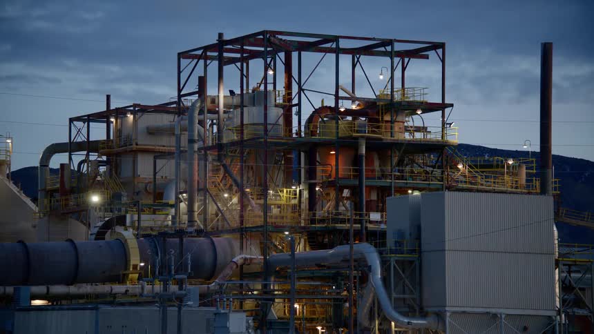 Lights glow on a factory or energy plant that is surrounded by a fence and stands near hills and brush