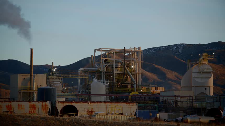 A refinery with a smokestack billowing smoke sits in a rural area
