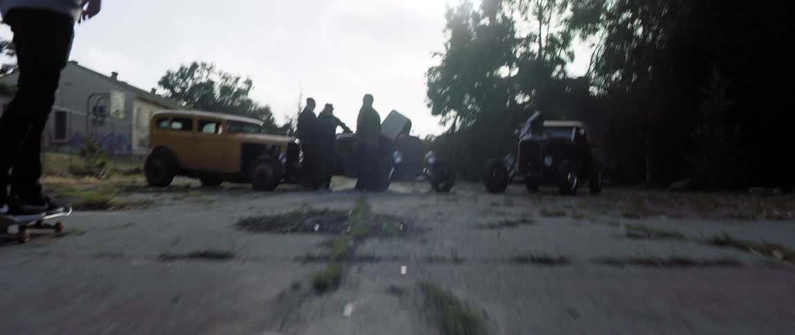A pair of skateboarders skate down a dilapidated road towards a group of men standing near vintage cars