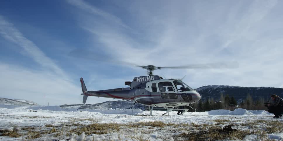 Two men in a field outside of a helicopter with its blades spinning