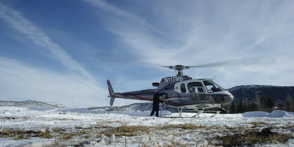 Helicopters sits on the snowy land with its blades spinning as someone wipes it down