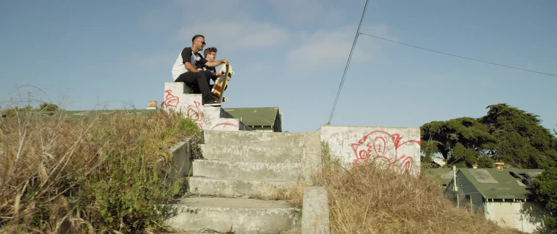 Two friends sit and hold skateboards on an outdoor staircase overlooking a neighborhood