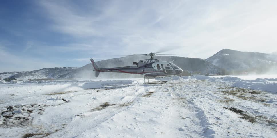 A helicopter hovers over a snow covered field near mountains