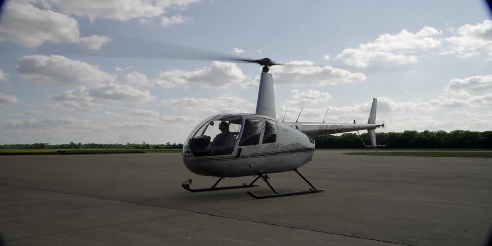 A helicopter sits on a pavement with its rotors still spinning
