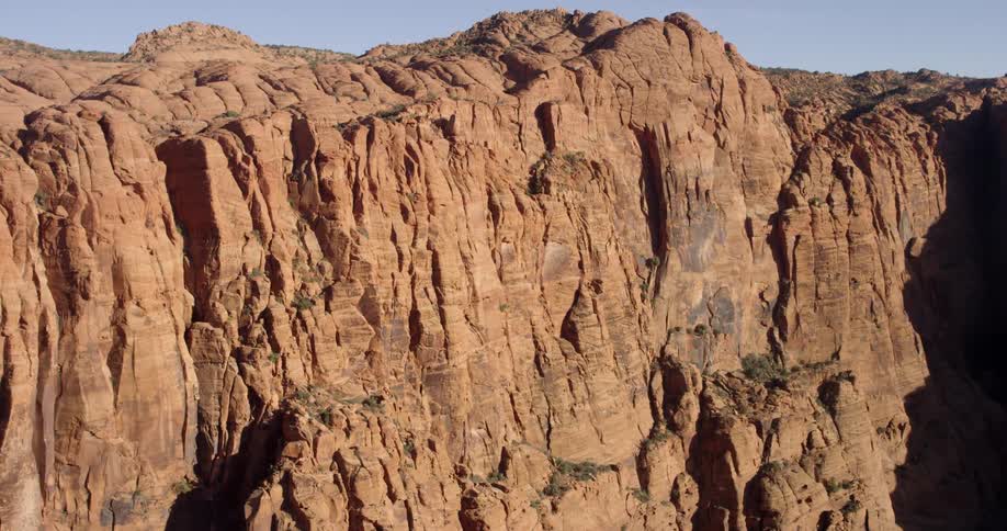 Sheer cliff of red striated stone borders scrub topped area under clear sky