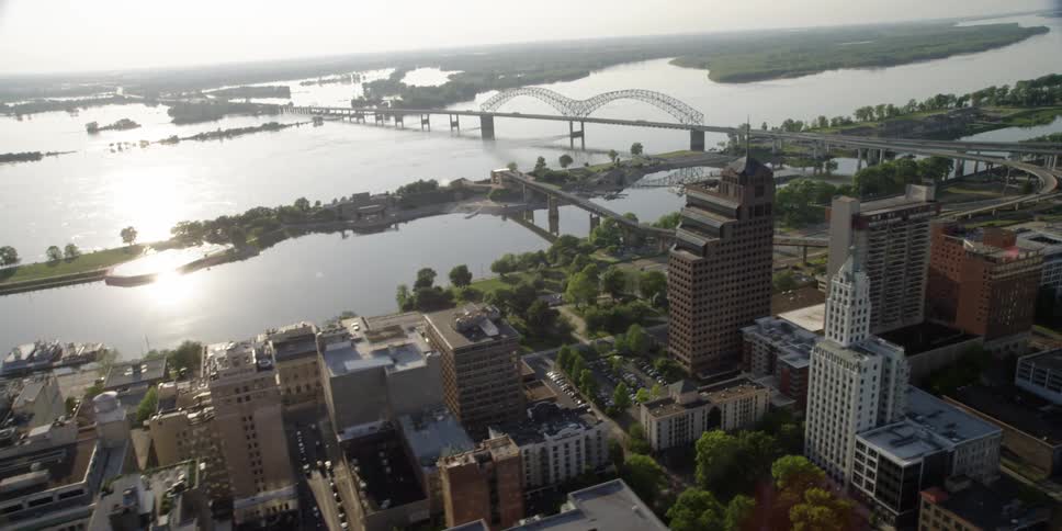 Passing over the rooftops and tree tops of a city with the sun shining and reflecting off the surface of the water