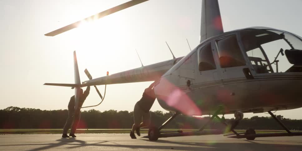 Two men tilt and push a helicopter into position on a rural airstrip on a bright sunny day