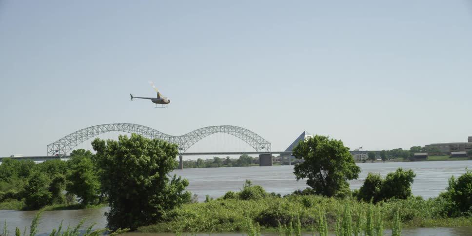 A helicopter flies over a river and toward a small city