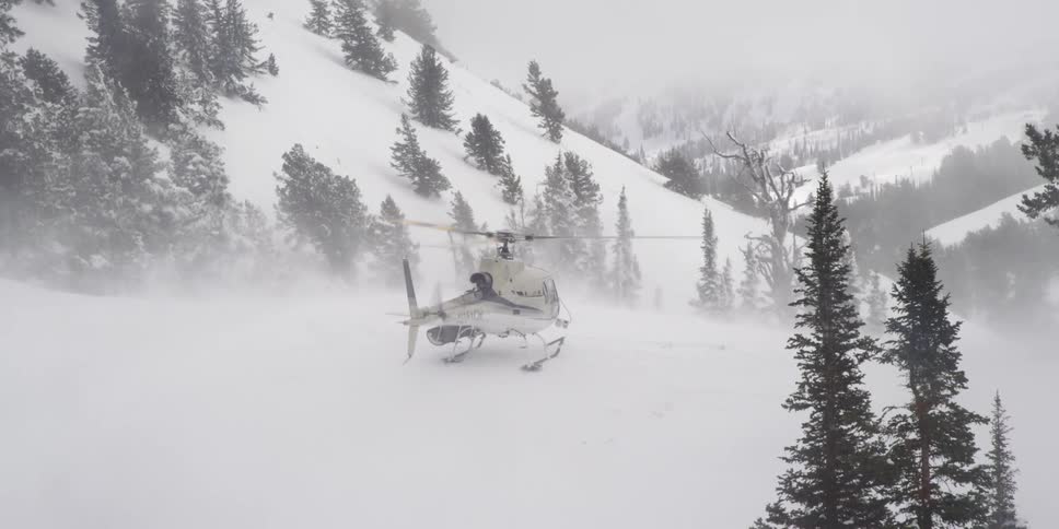 A helicopter sits on the snow in a nature area with its blades spinning