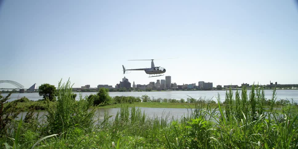 Lush green grass and plants on shore in the wind as a helicopter flies past with the city skyline on the horizon