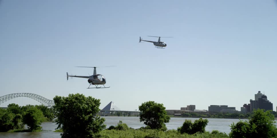 Two helicopters flying in the sky with a bridge and city skyline in the background