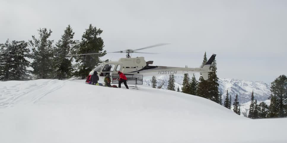 Helicopter sits on a snowy hill as its blades spin and a person in a red jacket is nearby