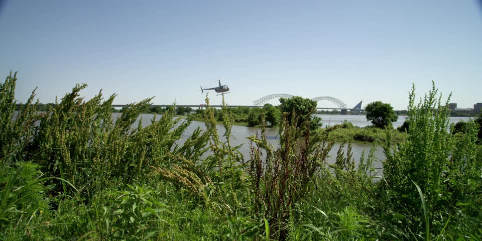 A helicopter flies over a river near a bridge and a city