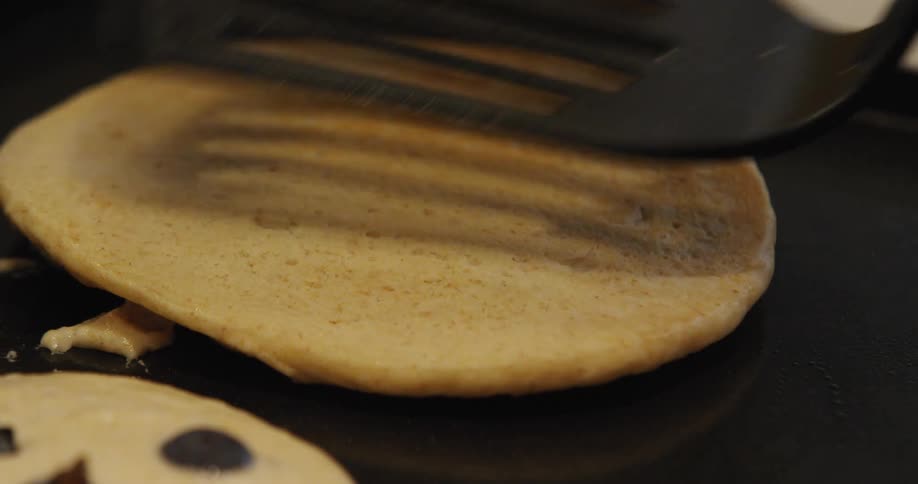 Chocolate chip pancake cooking on a griddle is flipped over with a plastic spatula