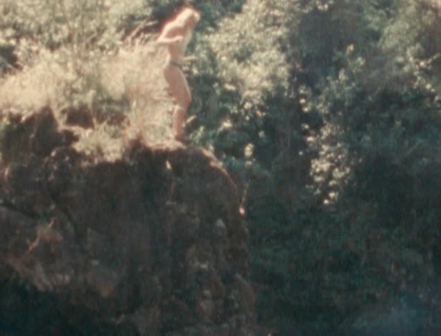 A young woman wearing a bikini jumps from a rocky ledge into a lagoon in a forest