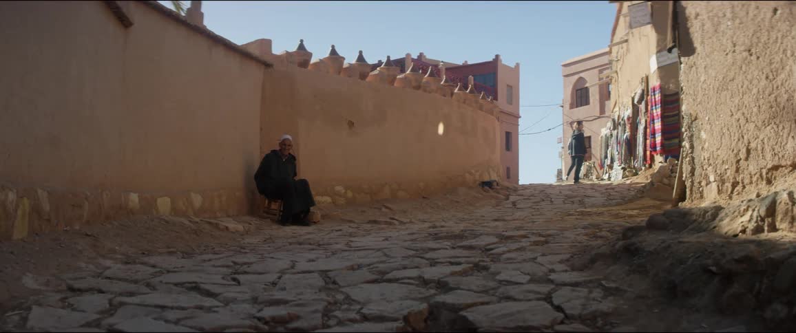 An elderly man sitting in the shadow of a stone wall in the alleyway of a town of stone buildings