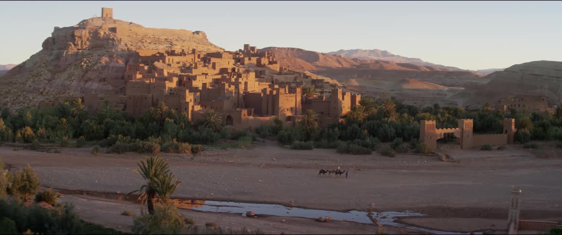 A man and several camels travel past an ancient city