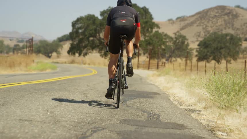 A bike rider pedals on an empty rural road with hills on the horizon on a bright sunny day