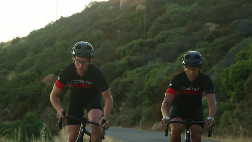 Two men race on bicycles down a rural road near a hilside