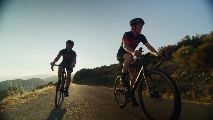 Two men ride bicycles down a narrow road surrounded by hills